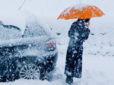 Person standing in the snow under an umbrella next to the back of a black car