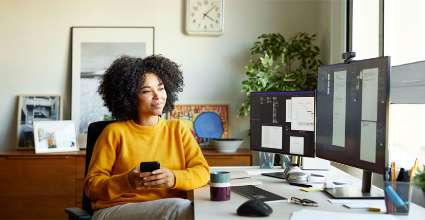 woman in a gold top holding a cell phone, looking at computer screens